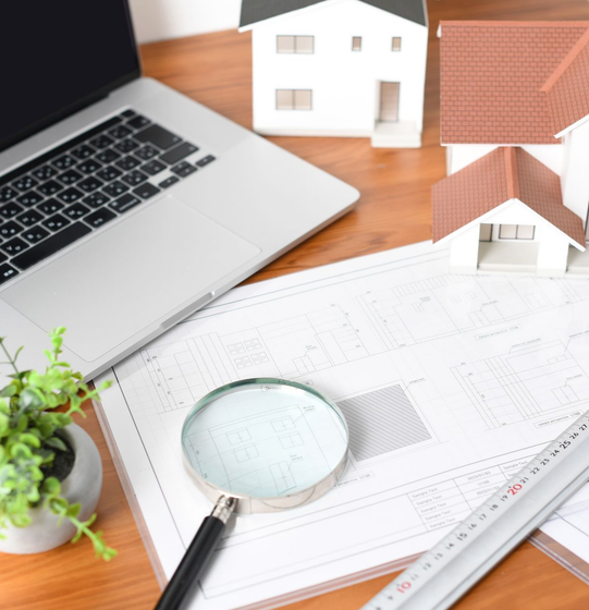 Ordinateur portable, plans, loupe et modèles de maisons sur un bureau, probablement pour la planification immobilière ou architecturale.