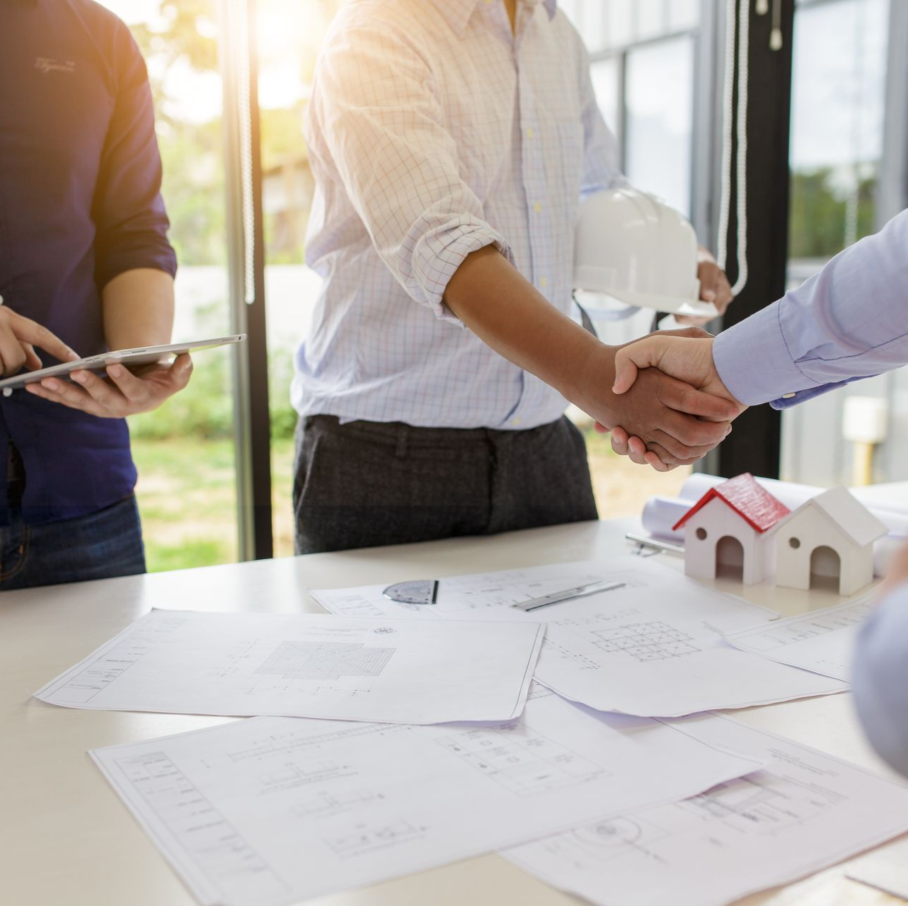 Des gens se serrant la main autour d'une table de plans de construction, de modèles de maisons et d'une tablette dans un bureau.
