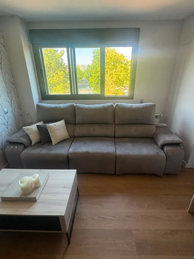 Sala de estar moderna con sofá gris, mesa de centro y gran ventanal con vistas a árboles verdes.