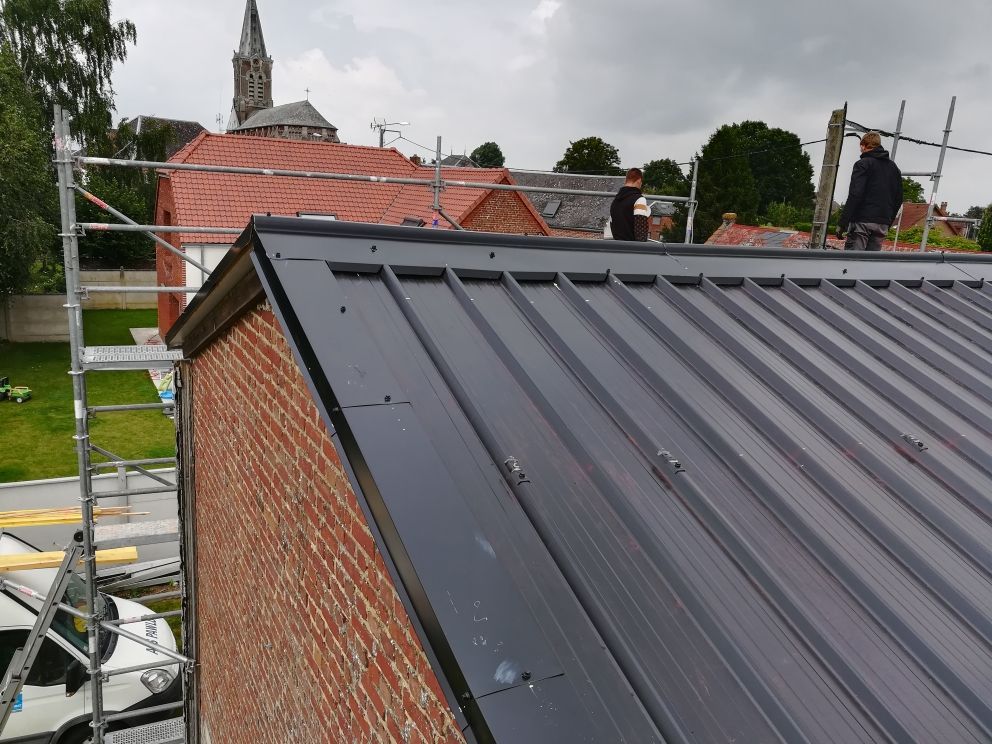 Des ouvriers installent un panneau de toiture métallique sur le toit d'un bâtiment en briques, à côté d'un échafaudage.