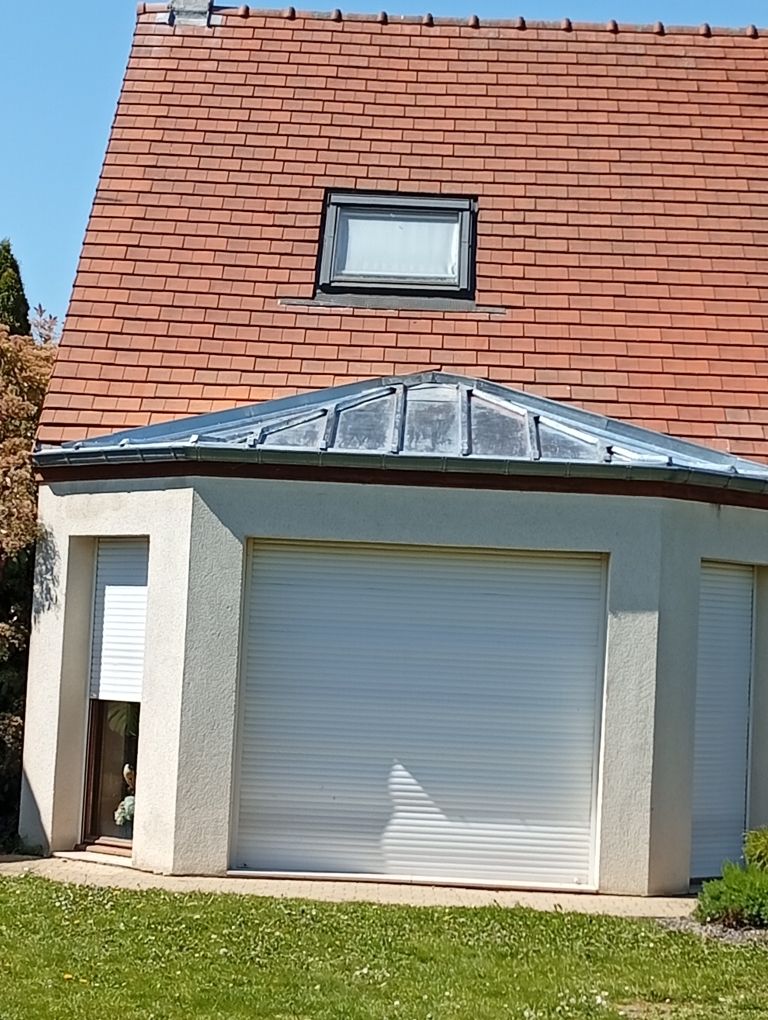 Extérieur d'une petite maison avec toit de tuiles, porte de garage et puits de lumière au-dessus de l'entrée.