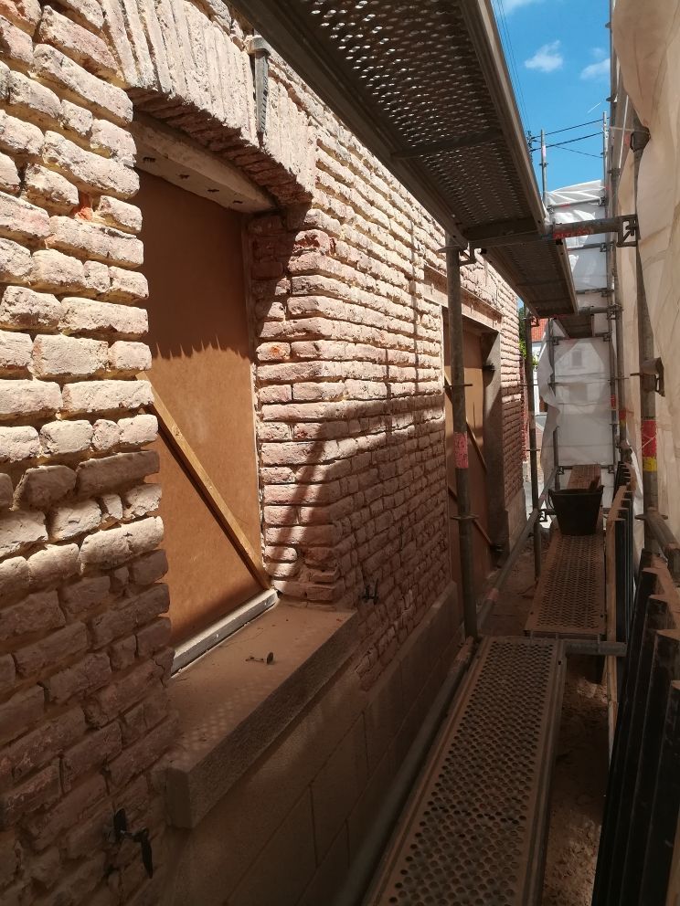 Ruelle étroite en briques avec un passage piétonnier en grille métallique et une fenêtre fermée baignée de soleil.