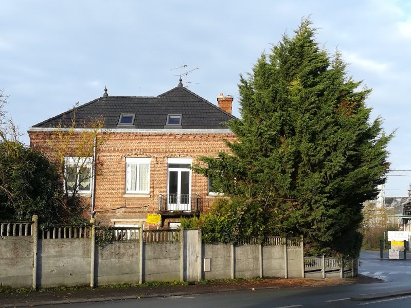 Maison en briques au toit sombre, derrière une clôture et un grand arbre en bord de route.