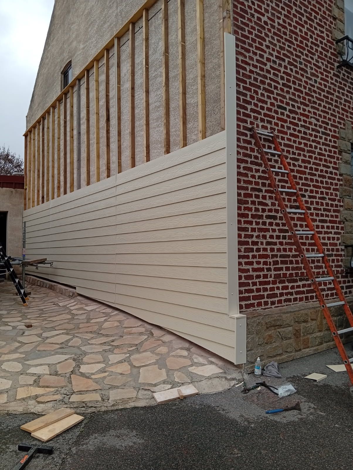 Mur extérieur en construction avec bardage, montants apparents et angle en briques à côté d'une allée en pierre.