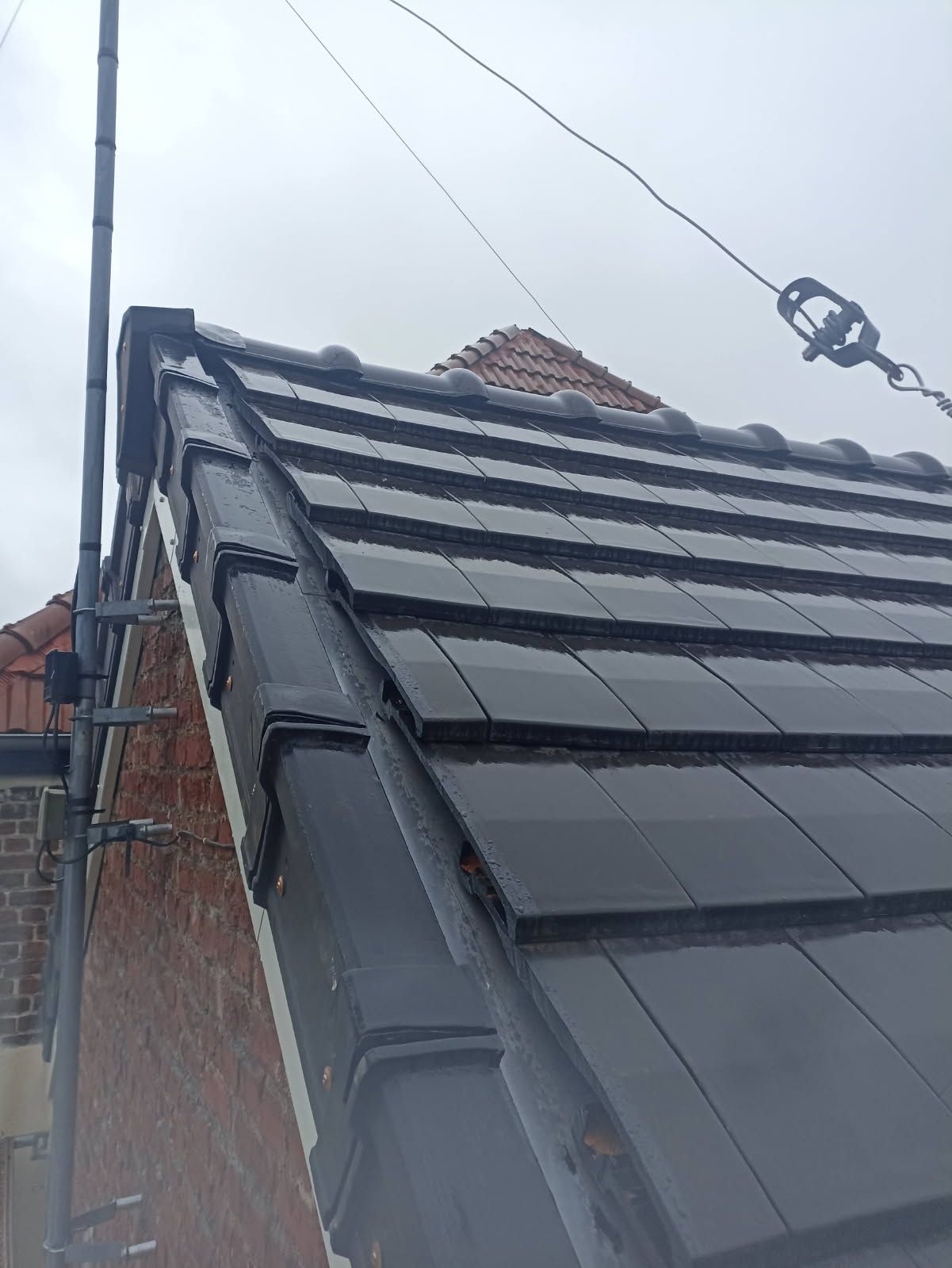 Gros plan sur un toit pentu de tuiles avec gouttières, à côté d'une maison en briques et de lignes électriques au-dessus.