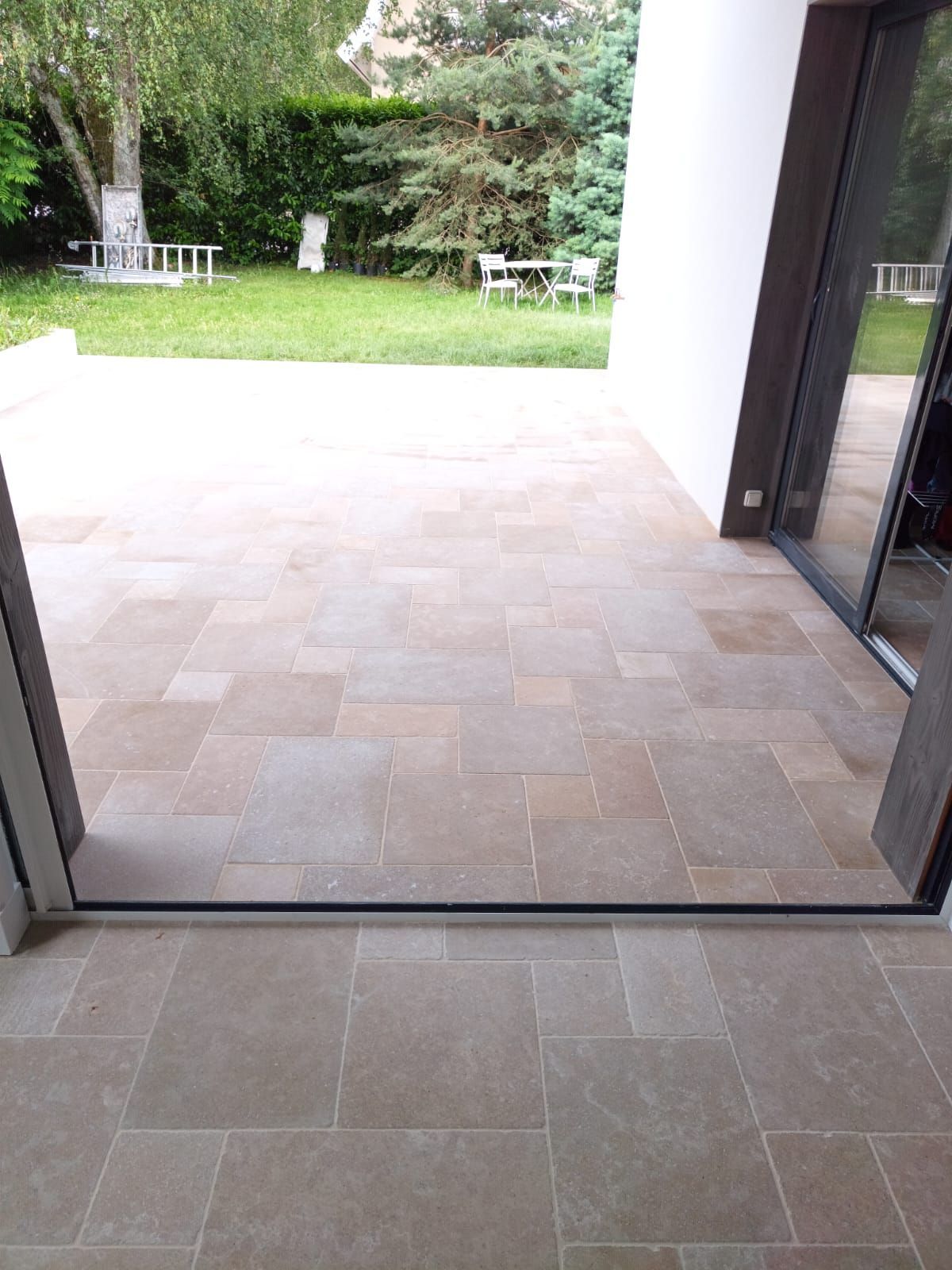 Une terrasse carrelée donne sur une cour gazonnée et arborée, visible depuis une porte vitrée entrouverte.