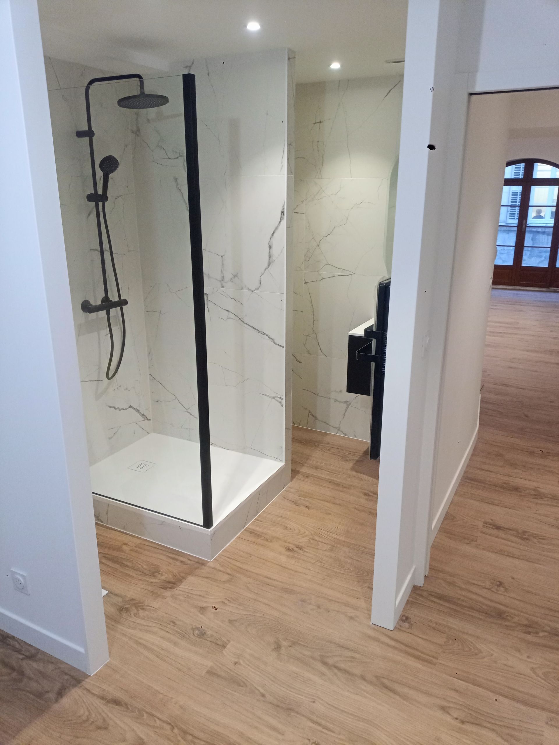 Salle de bains moderne avec douche à l'italienne. Robinetterie noire et murs imitation marbre. Sol en bois.