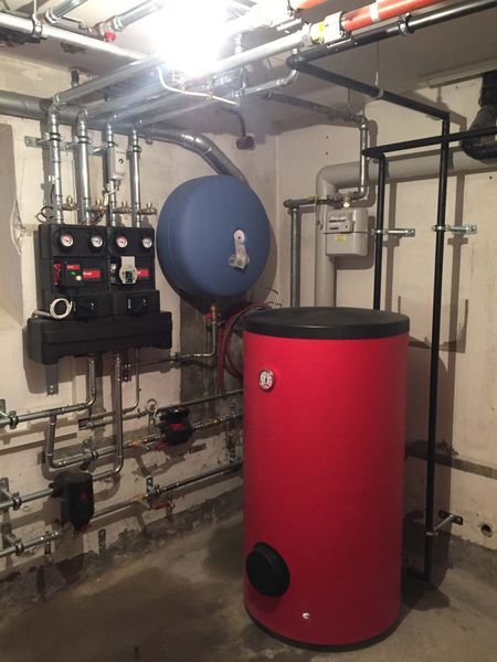 Heizraum mit rotem Warmwasserspeicher, blauem Ausdehnungsgefäss, Rohrleitungen und Bedienfeld.