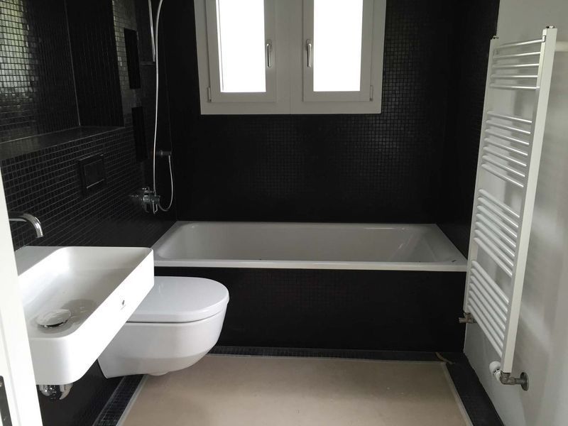 Modernes Badezimmer mit schwarz gefliesten Wänden, weisser Badewanne, Toilette, Waschbecken und Handtuchheizkörper in Fensternähe