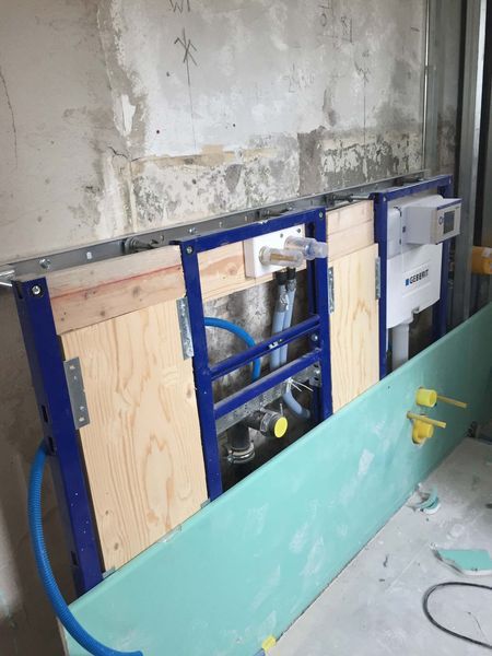Bauarbeiten an einer Wand mit blauem Rahmen, freiliegenden Rohren und einer darunter lehnenden grünen Platte.