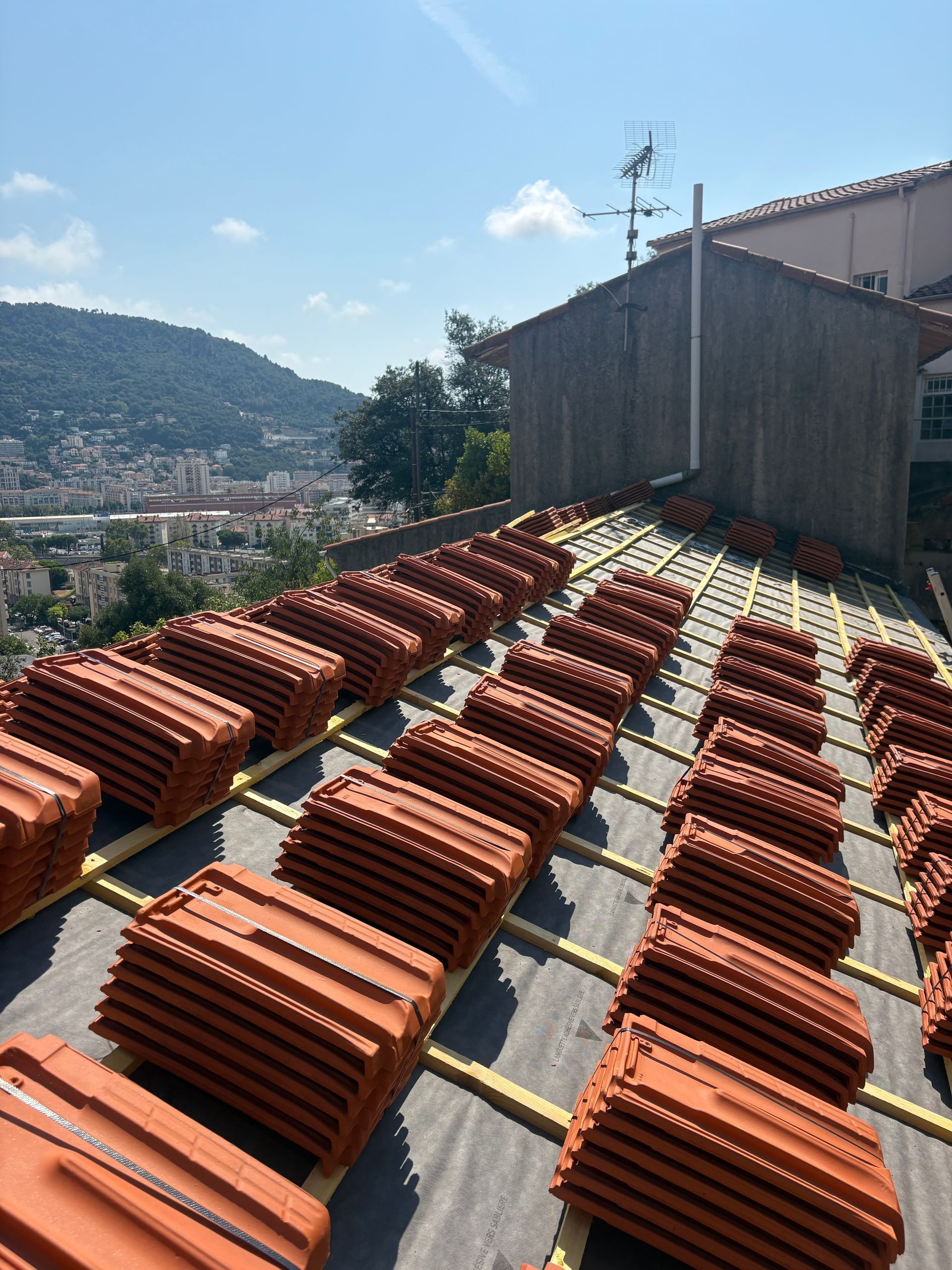 Des piles de tuiles en terre cuite brun rougeâtre disposées sur un toit en pente, avec une vue sur la ville en arrière-plan.