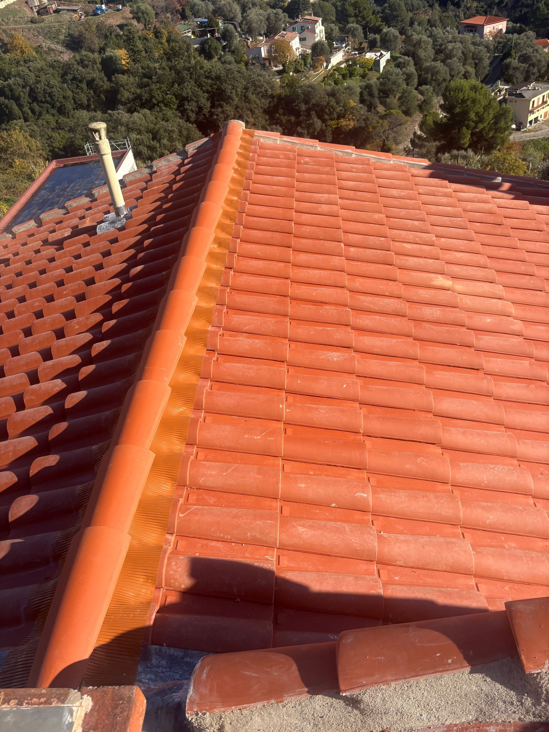 Vue aérienne d'un toit de tuiles en terre cuite avec une arête centrale, montrant un conduit de ventilation et un arrière-plan arboré.