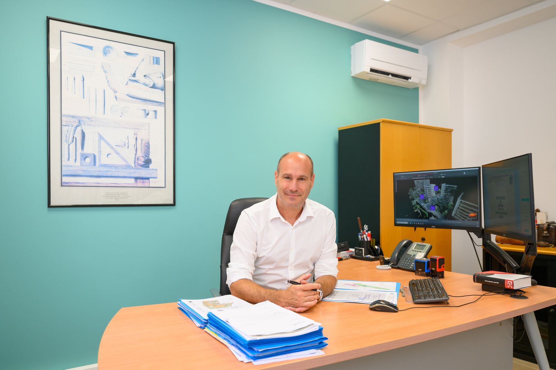 Un homme en chemise blanche est assis à un bureau dans un bureau turquoise, avec des écrans d'ordinateur, des papiers et une affiche murale.