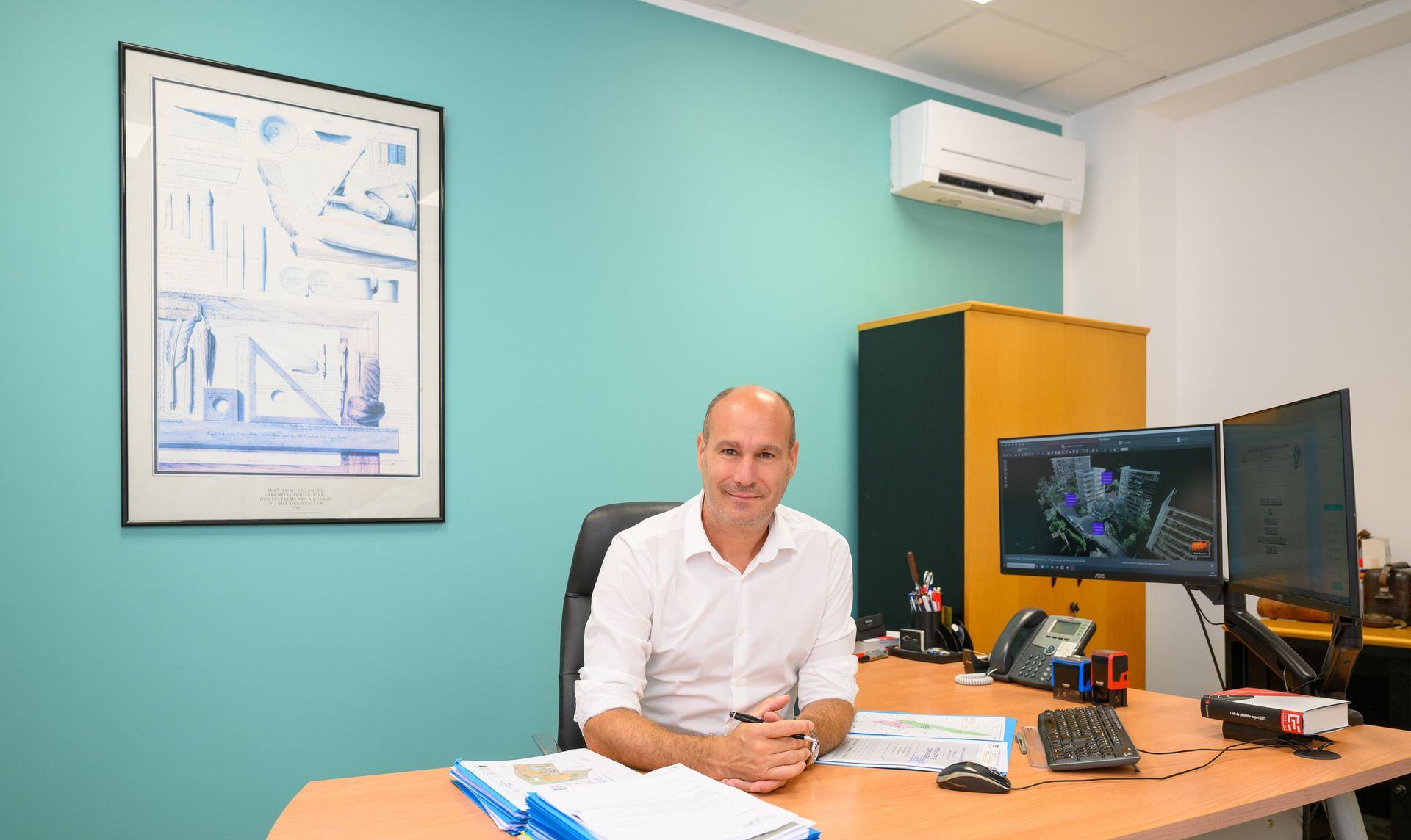 Une personne assise à un bureau dans un bureau aux murs turquoise, avec des écrans d'ordinateur et des graphiques encadrés.