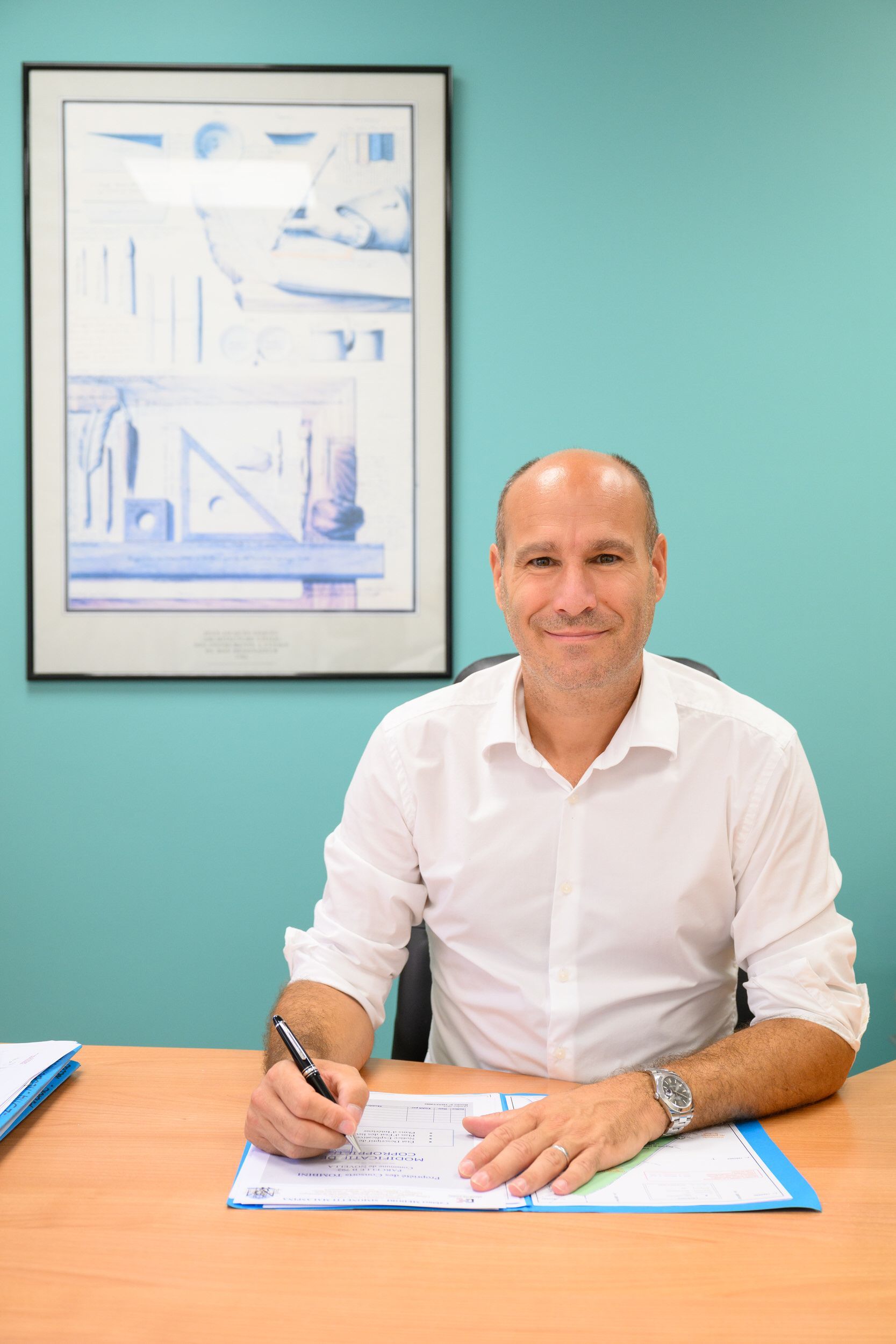 Un homme en chemise blanche signe des documents à un bureau dans un bureau aux murs turquoise.