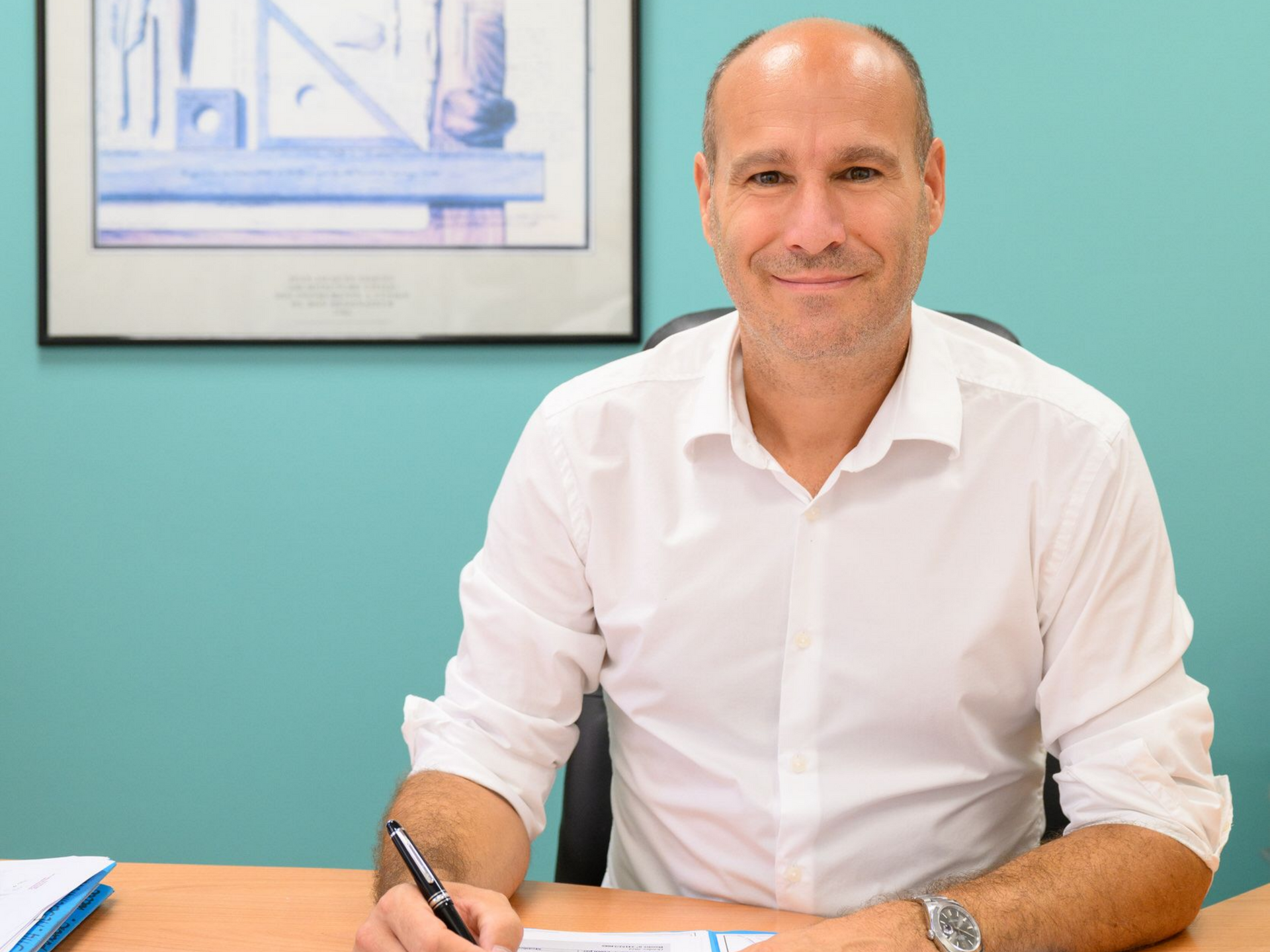 Un homme en chemise blanche, assis à un bureau, sourit dans un bureau bleu turquoise, tenant un stylo.