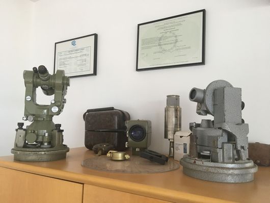 Étagère d'instruments d'arpentage anciens sur une table en bois, sous des certificats encadrés.