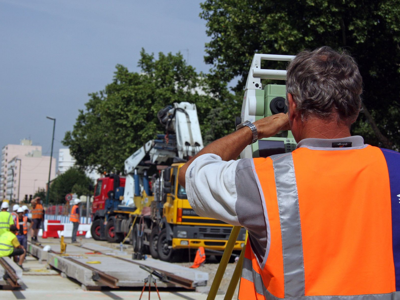 Une équipe de construction dirige des travaux routiers à l'aide de grues et de camions ; un ouvrier portant un gilet de sécurité orange se trouve au premier plan.