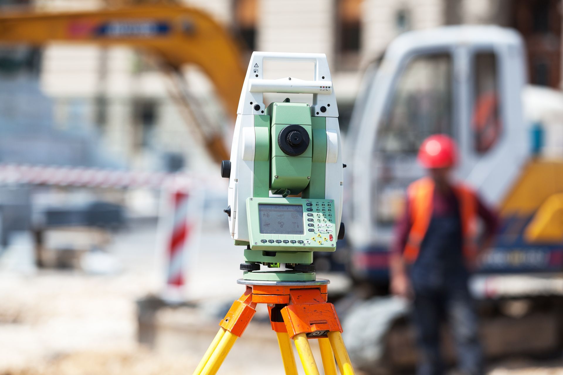 Instrument topographique sur trépied sur un chantier routier, avec un ouvrier et une excavatrice flous en arrière-plan.