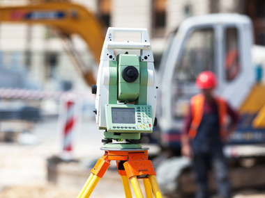 Instrument de topographie vert sur trépied orange sur un chantier de construction, avec des ouvriers et des machines flous en arrière-plan.
