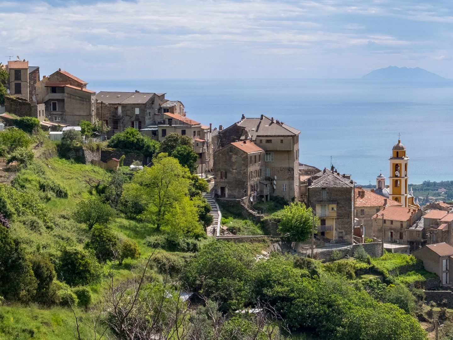 Maisons en pierre perchées sur une colline dominant un versant verdoyant surplombant une mer bleue calme et une île au loin.