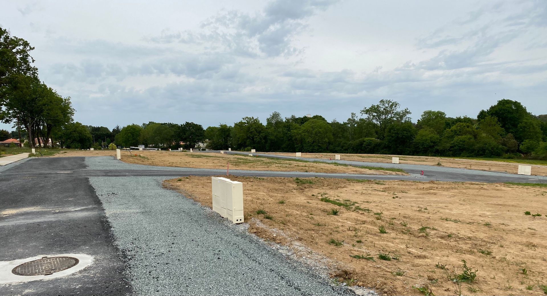 Chemin de gravier et chantier désert sous un ciel nuageux, avec des arbres en arrière-plan.