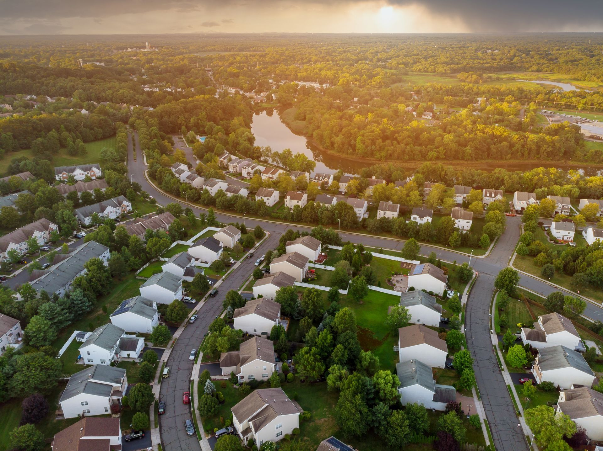 Vue aérienne d'un quartier résidentiel avec des rues sinueuses, des maisons, des arbres et un étang au coucher du soleil.