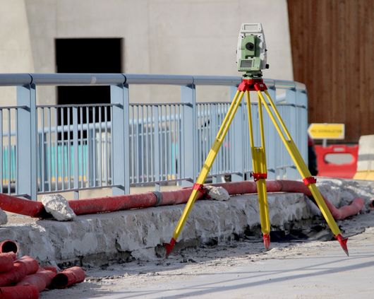 Trépied de topographie avec niveau laser sur un chantier de construction à côté d'un trottoir et d'une rambarde.