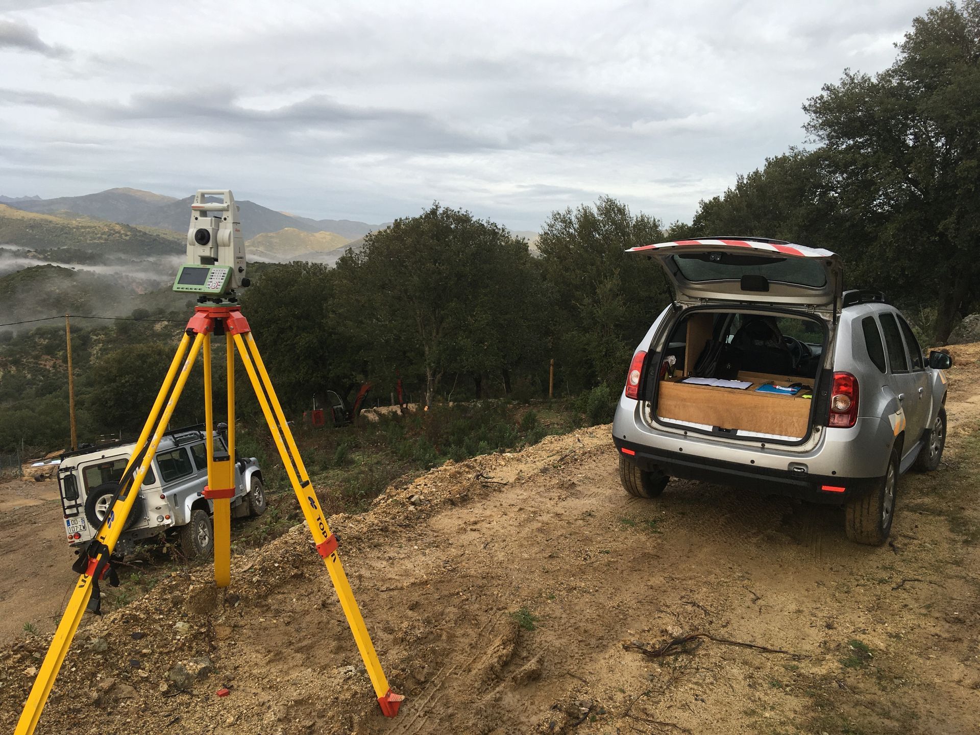 Du matériel topographique sur trépied se trouve à côté d'un SUV argenté à hayon ouvert, sur un chemin de terre dans une zone rurale vallonnée.