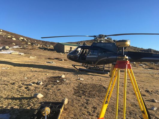 Un hélicoptère noir sur un terrain rocailleux, à côté d'un instrument de topographie sur trépied jaune, dans un paysage désertique.