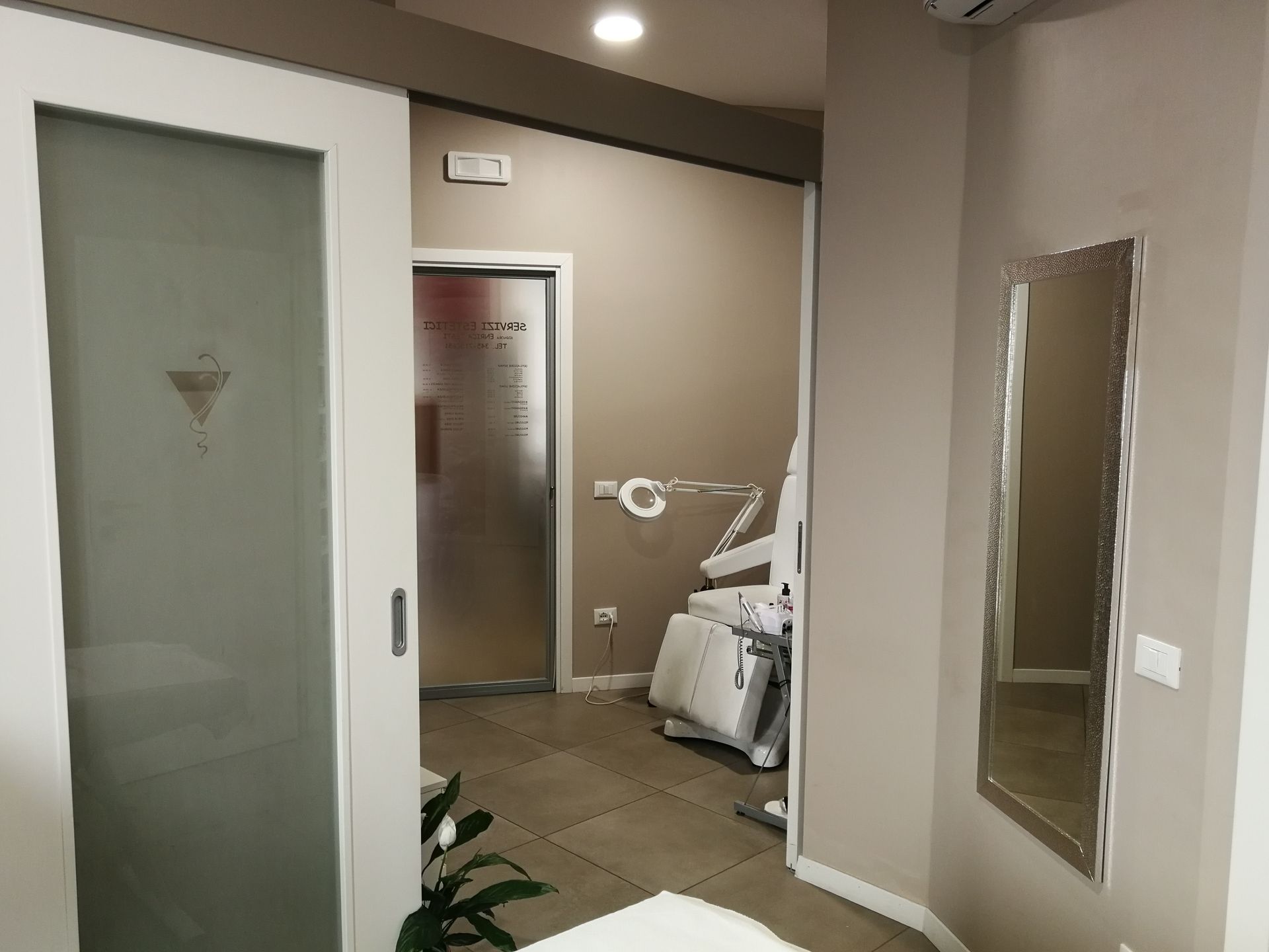 Interno della cabina estetica con porta in vetro smerigliato, specchio e attrezzature. Luci soffuse, colori neutri.