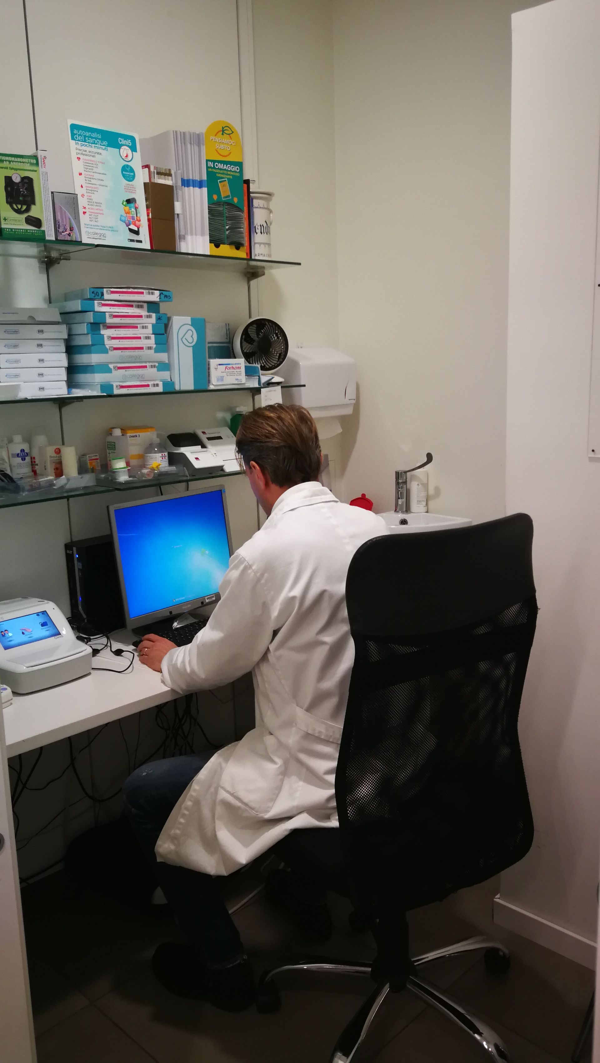 Uomo in camice seduto alla scrivania, mentre usa un computer. Sullo sfondo, scaffali con materiali.