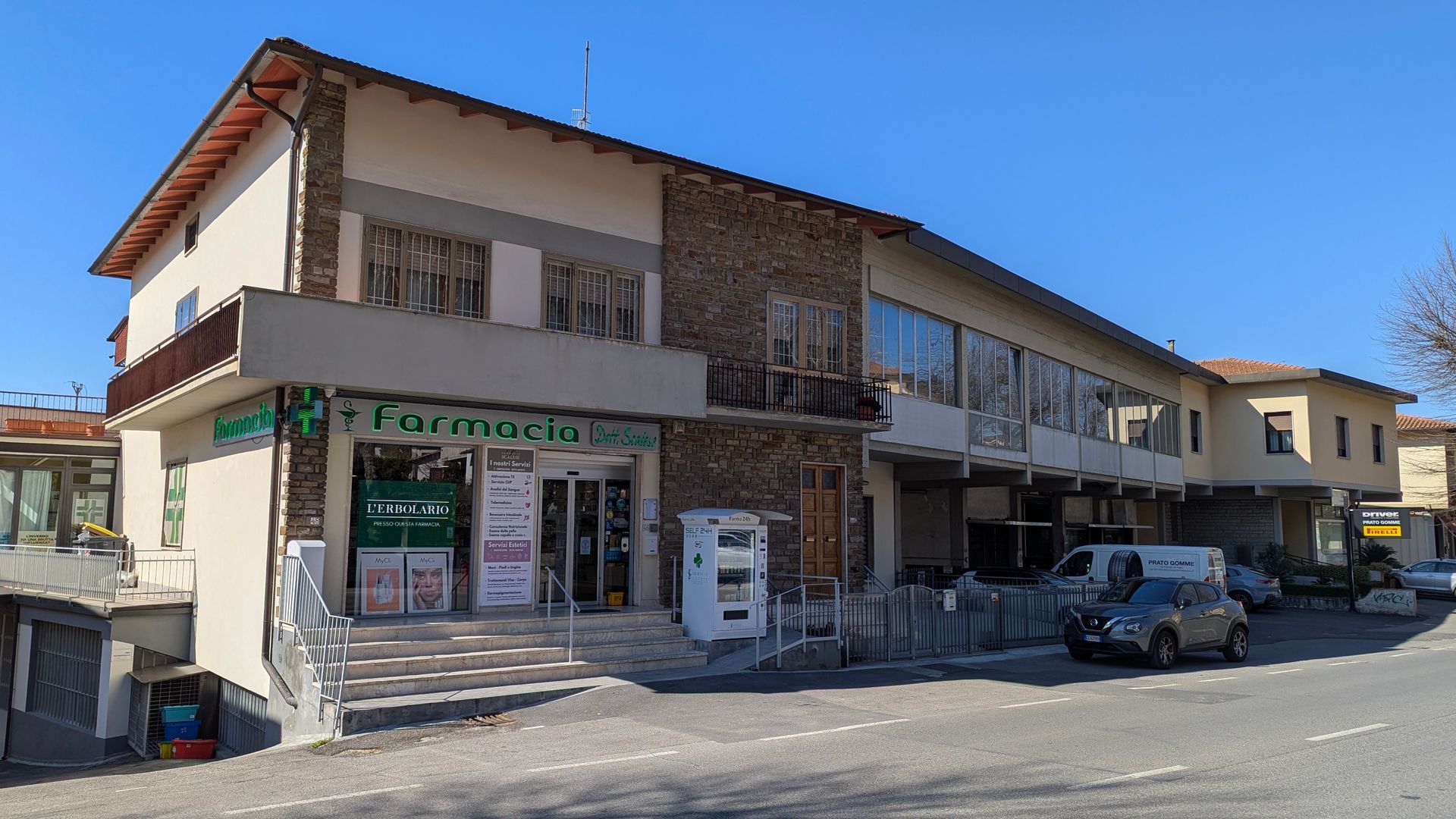 Un edificio a due piani con la farmacia, gradini e parcheggio nelle giornate di sole.