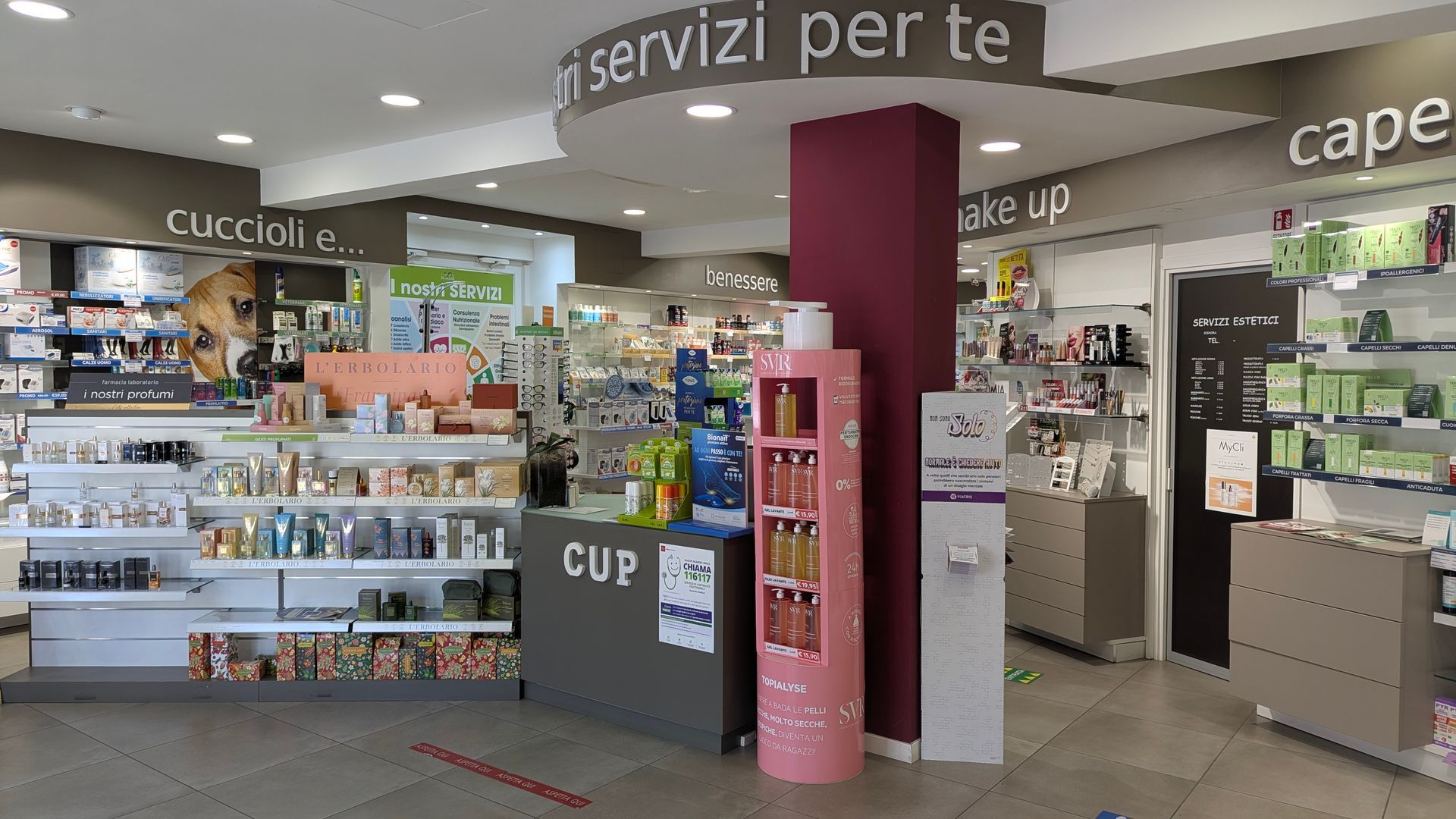 Interno della farmacia, con scaffali pieni di prodotti e un pilastro centrale.