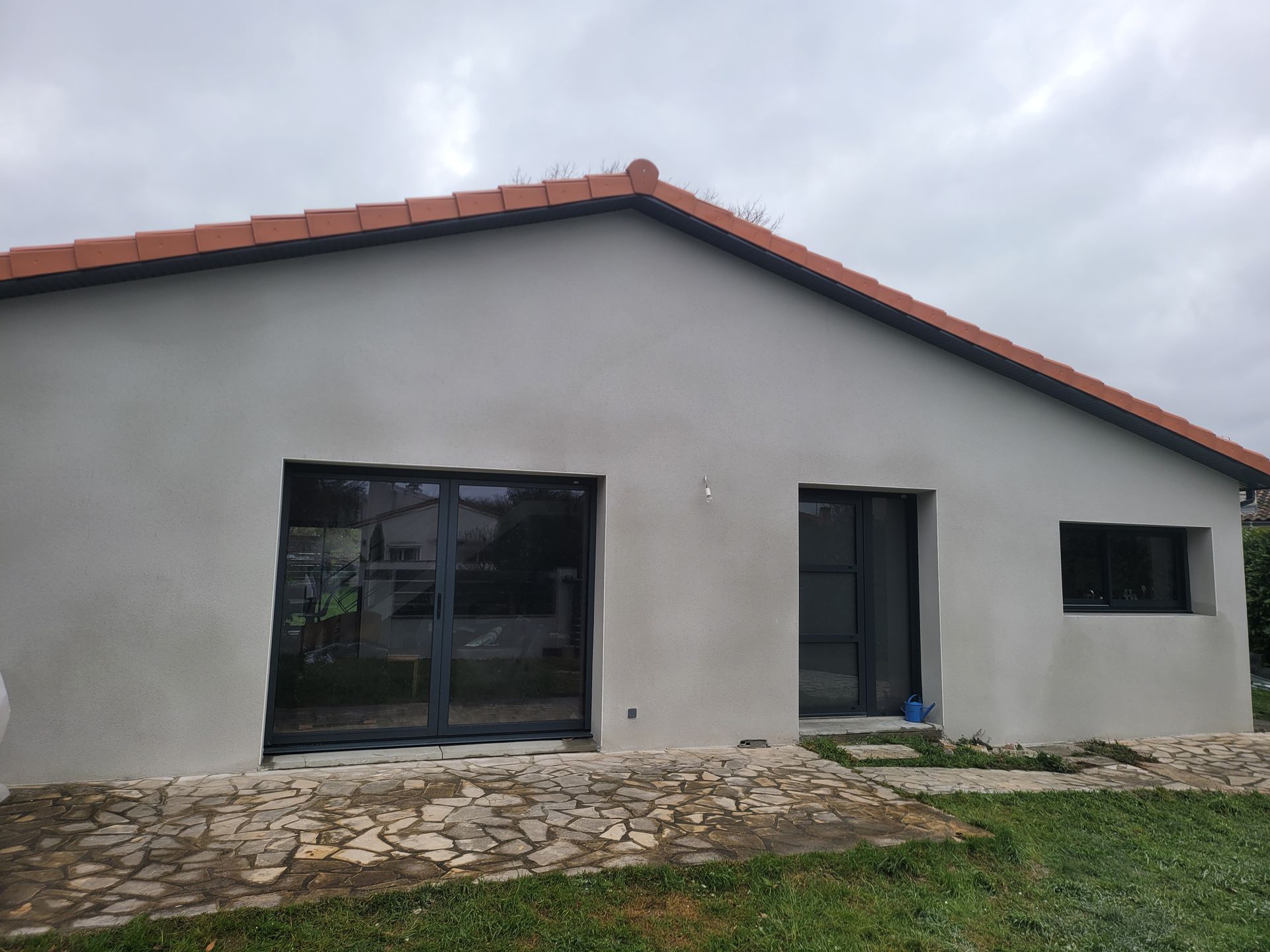 Maison grise aux fenêtres à encadrement sombre et au toit de tuiles rouges.