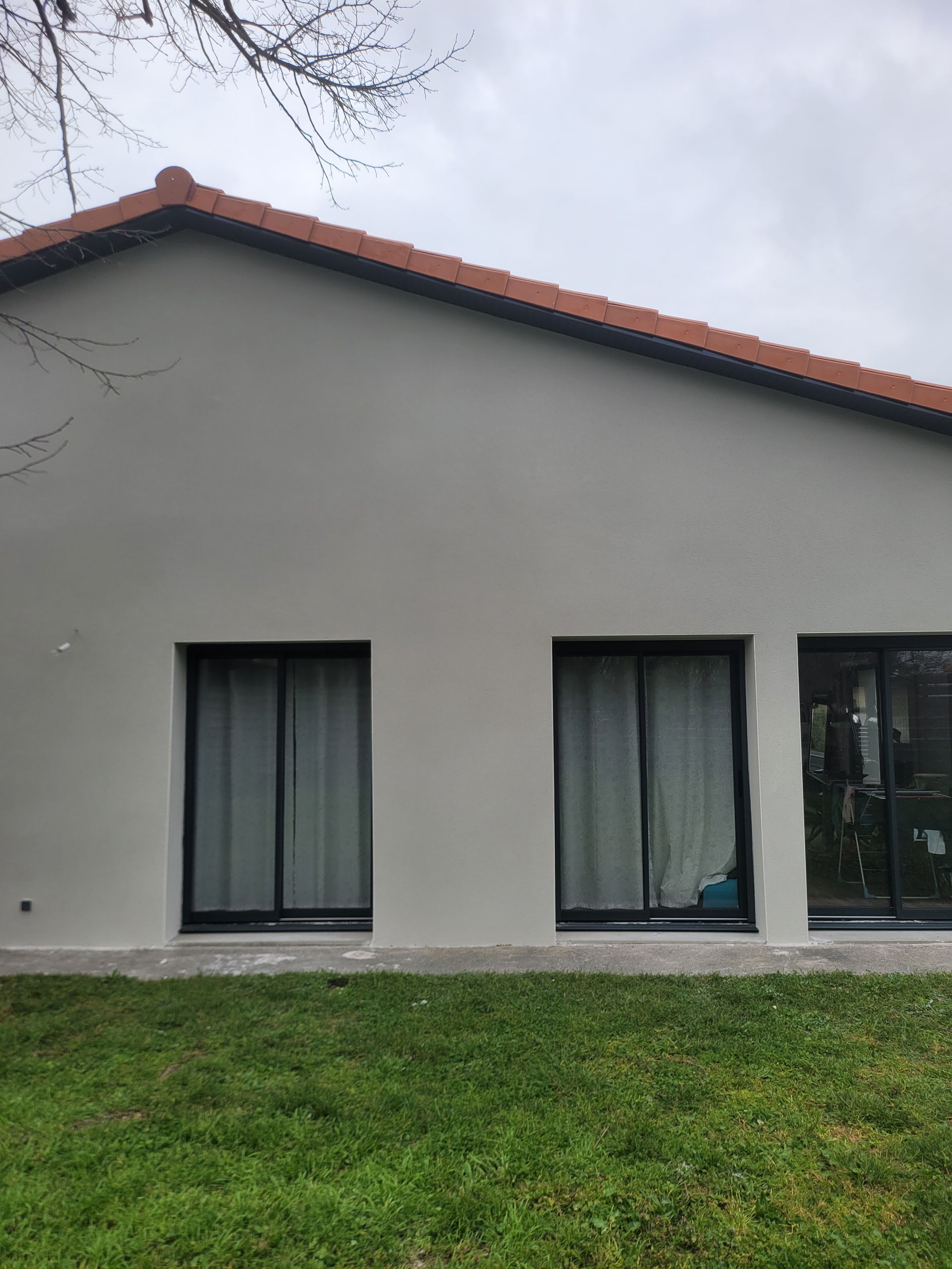 Maison grise à l'extérieur, avec trois fenêtres à cadre noir et un toit de tuiles rouges.