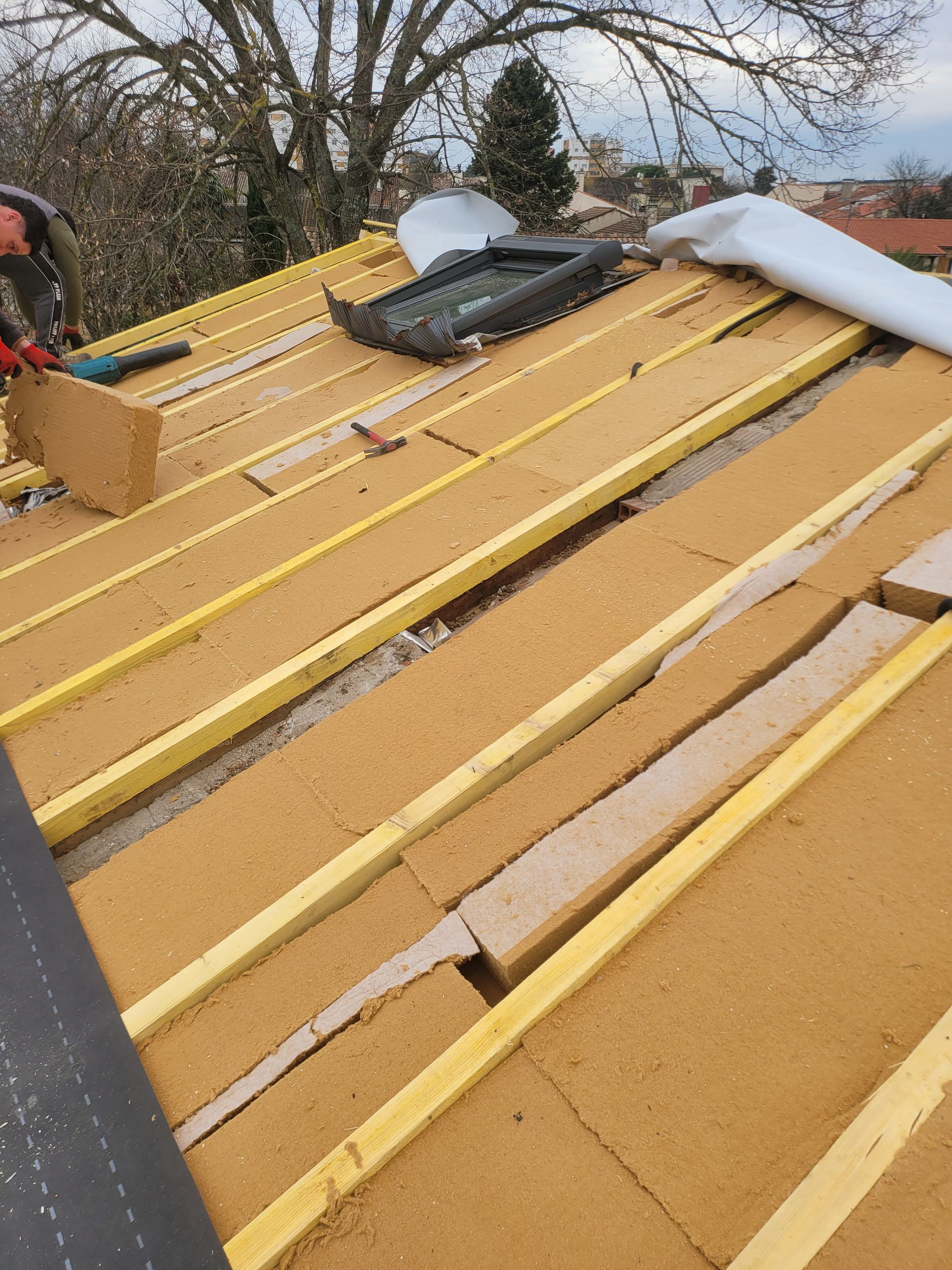 Un ouvrier du bâtiment installe de l'isolant sur un toit avec un puits de lumière ; planches et outils sont visibles.