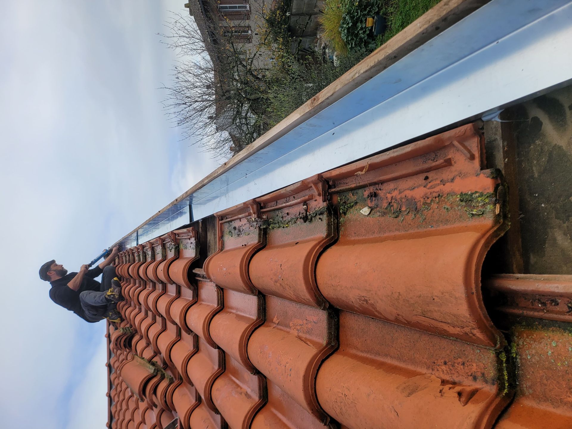 Un homme installe une gouttière sur un toit de tuiles. Le toit est brun rougeâtre et la gouttière argentée.