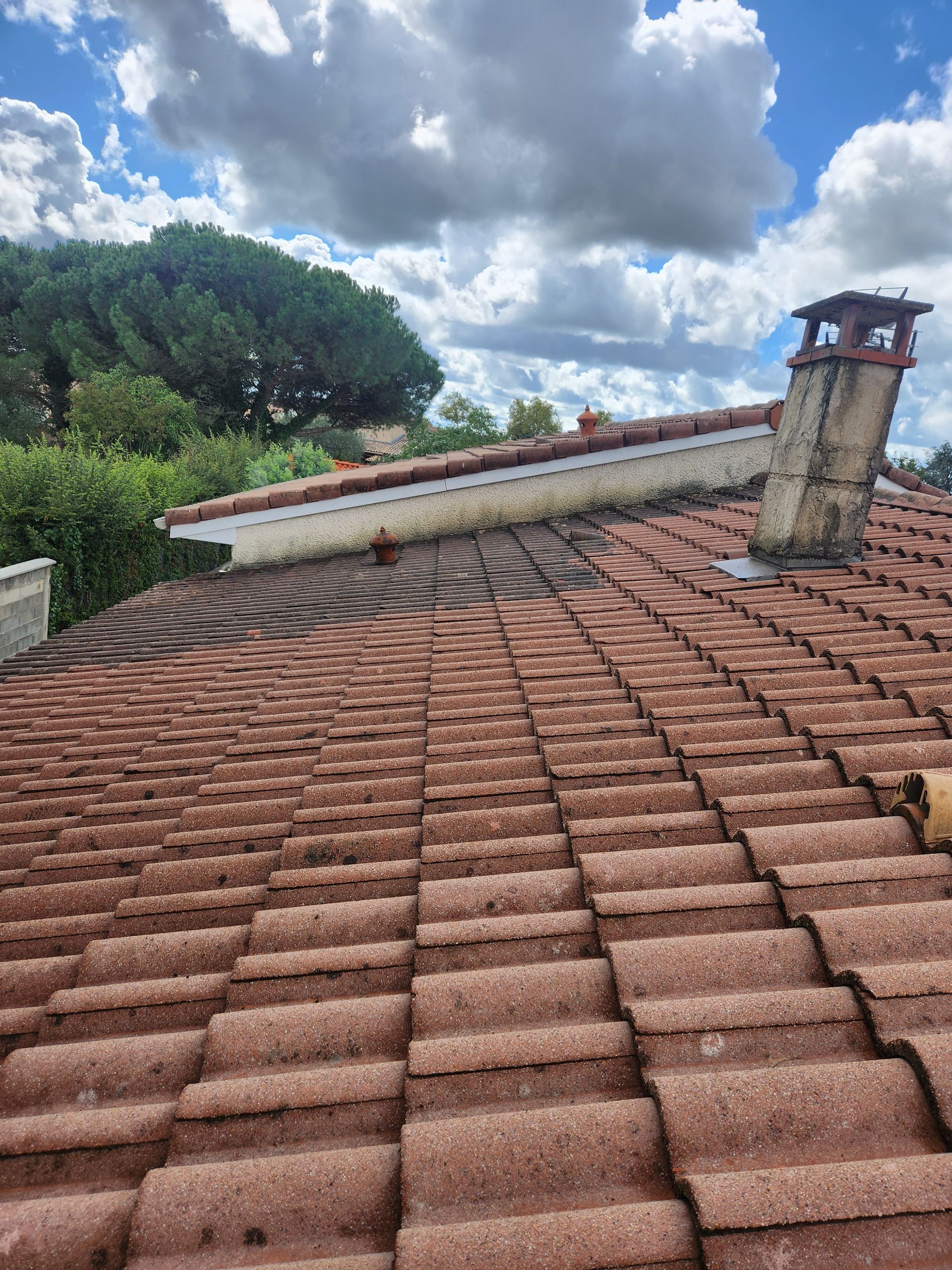 Toit de tuiles rouges avec cheminée se détachant sur un ciel bleu nuageux et des arbres verts.