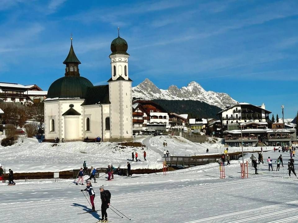 Verschneite Szene mit Kirche, Skifahrern und Bergkulisse.