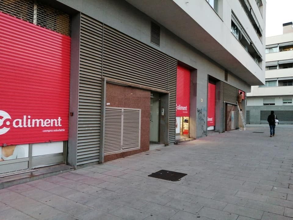 Tiendas a pie de calle con contraventanas rojas en una tranquila acera de la ciudad, una persona caminando a lo lejos.
