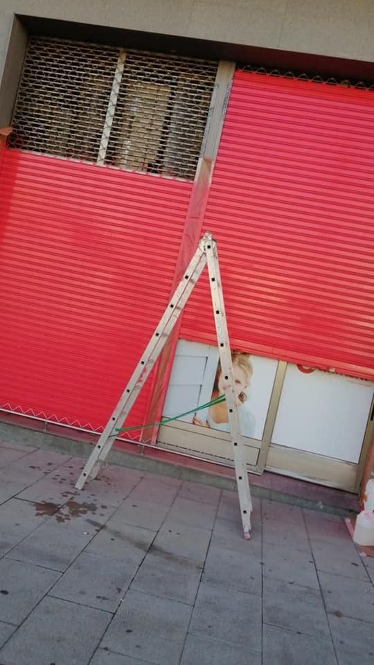 Una escalera se alza sobre una acera de baldosas frente a unas contraventanas rojas de una tienda y una ventana.