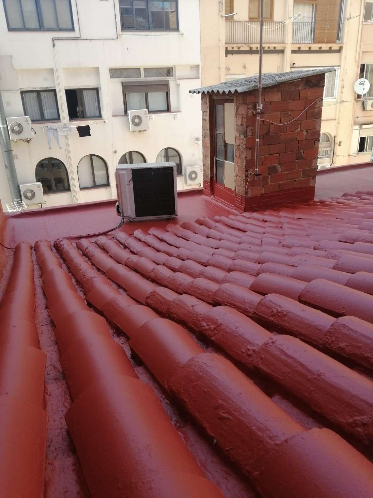 Tejado de chapa ondulada roja con una pequeña chimenea de ladrillo y edificios de apartamentos al fondo.