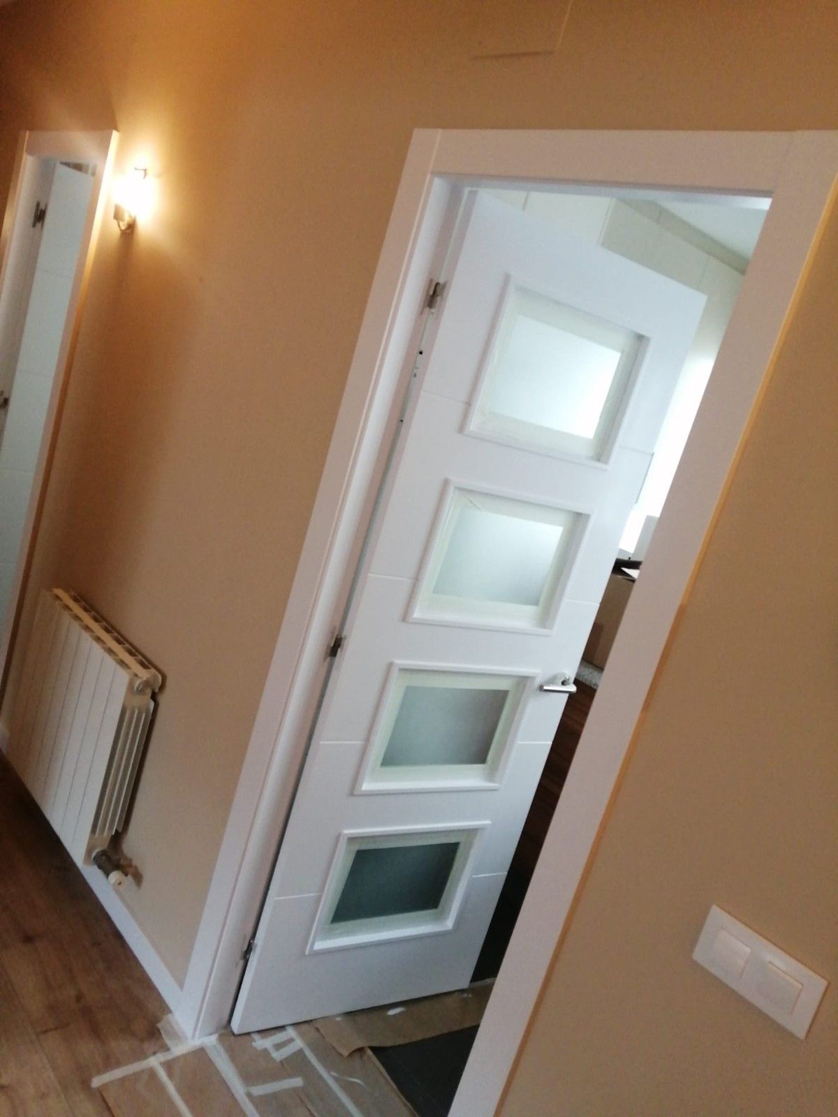 Vista inclinada de una puerta interior blanca abierta en un pasillo beige con un radiador de pared y un interruptor de luz.