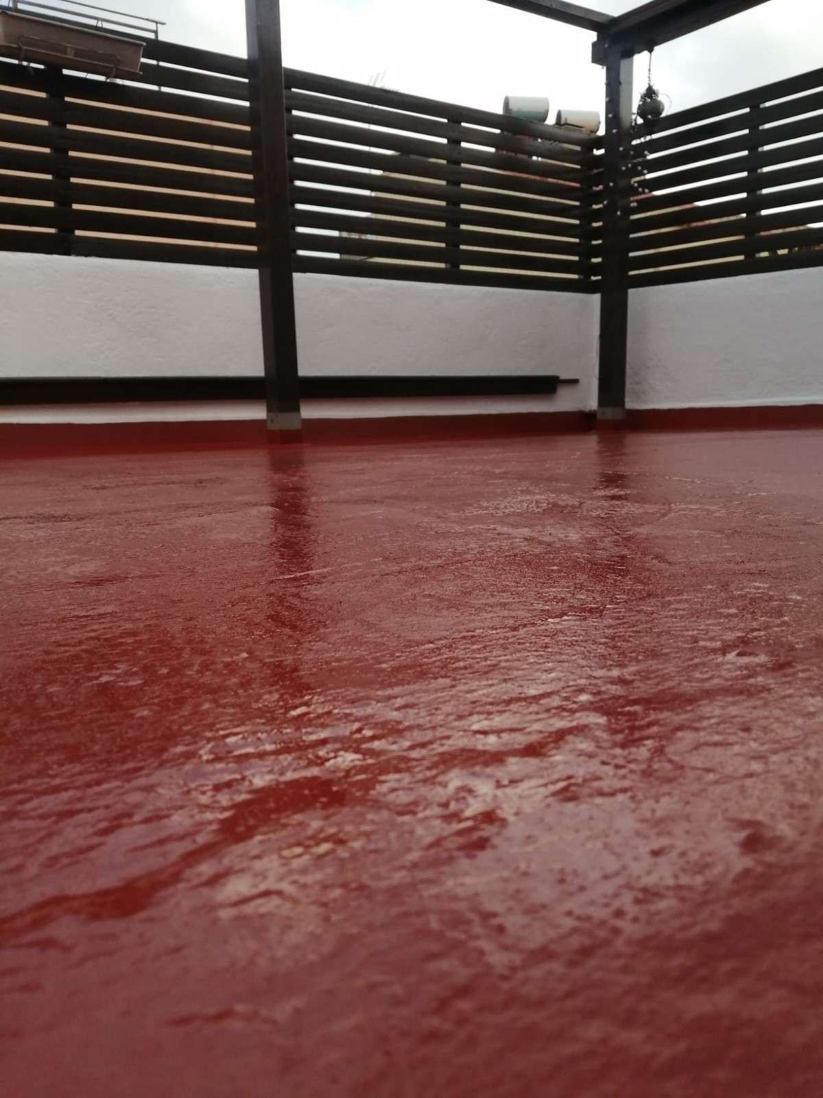 Suelo rojo y mojado en una terraza de azotea con barandilla de madera y postes grises bajo una luz nublada.