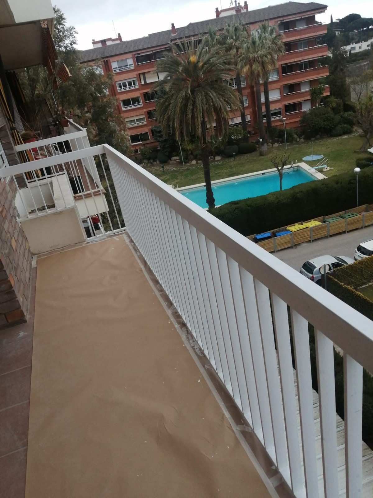 Balcón con vistas a una piscina en un patio, palmeras y un edificio de apartamentos bajo una luz nublada.