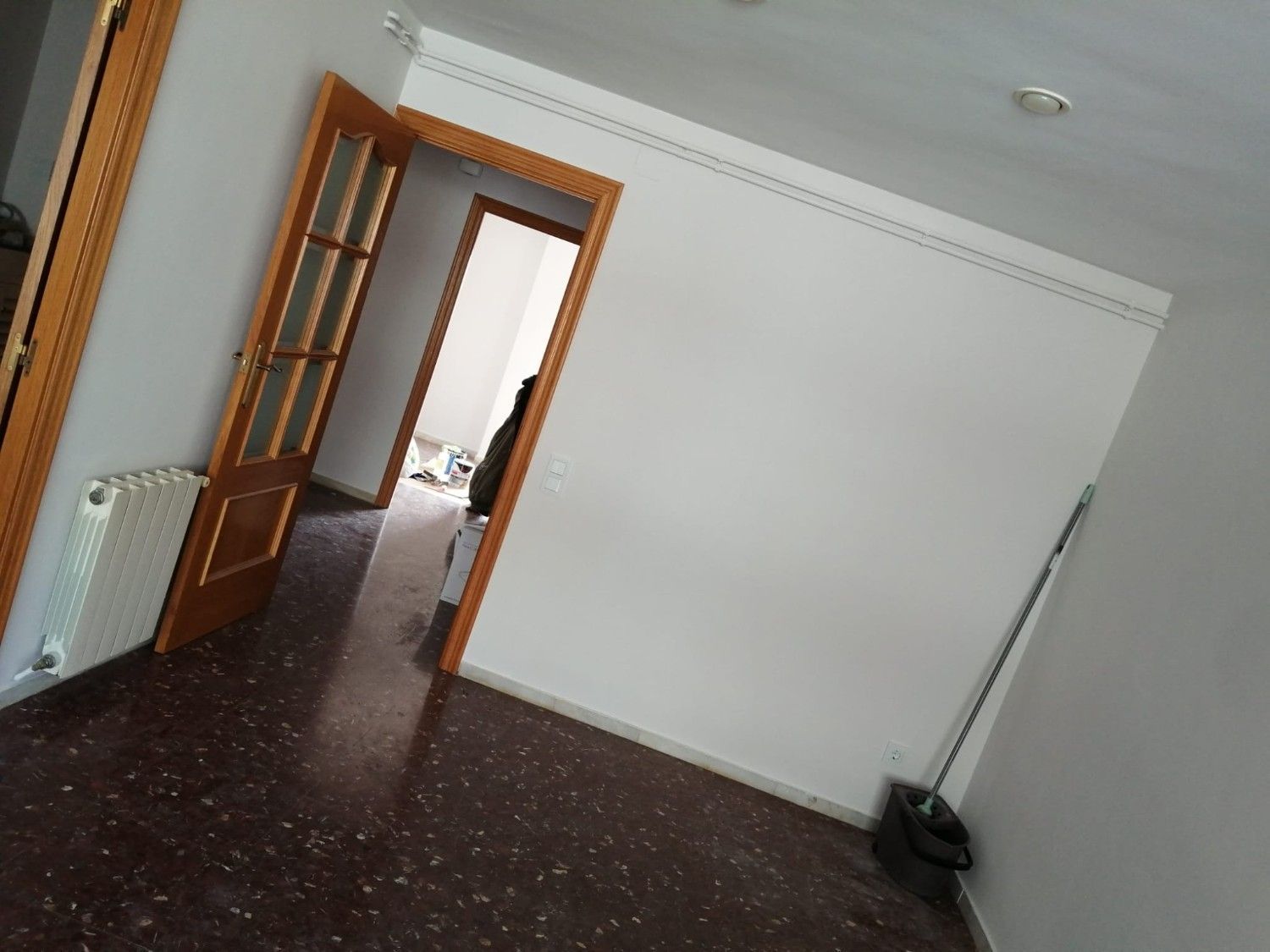 Habitación vacía con paredes blancas, suelo oscuro moteado, puertas de madera abiertas y una escoba en la esquina.