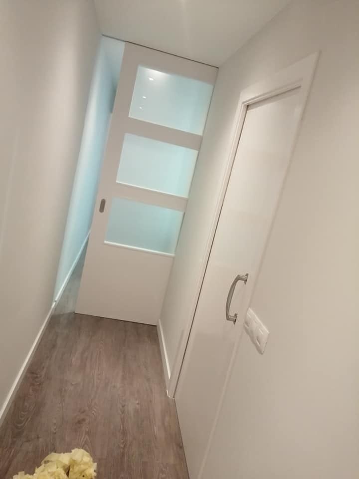 Pasillo estrecho con suelo de madera, paredes blancas, puerta de cristal esmerilado y una puerta interior blanca cerrada.