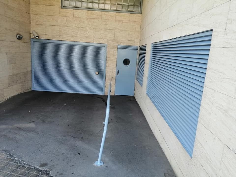 Área de estacionamiento techada con postes de barrera de línea blanca y puertas metálicas azules sobre paredes beige.