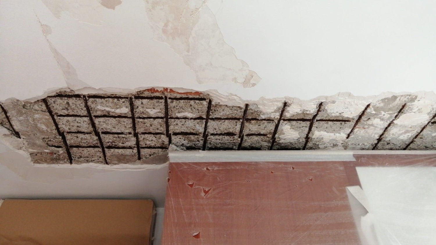 Pared de yeso dañada con malla metálica expuesta sobre una encimera y una caja de cartón.